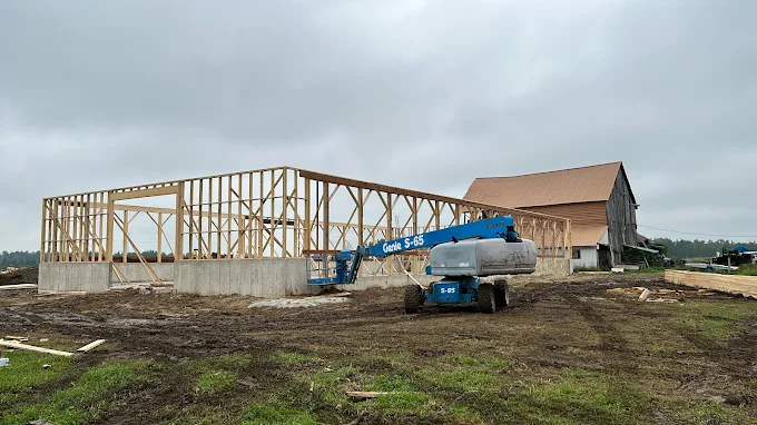 Post-frame building framing in progress beside a heritage barn